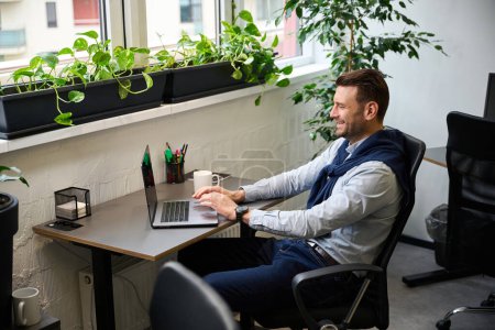 Foto de Sonriente hombre se comunica en línea utilizando un ordenador portátil, que trabaja en una cómoda oficina luminosa - Imagen libre de derechos