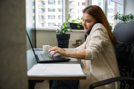 Foto de Hermosa mujer sentada en una silla de oficina con un teléfono móvil y un ordenador portátil, en el interior con muchas plantas de interior - Imagen libre de derechos