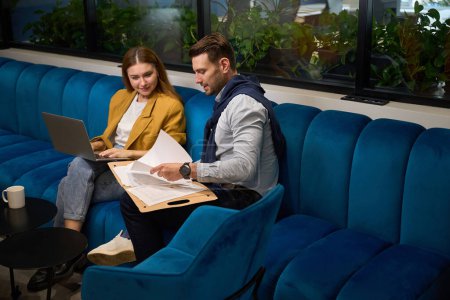 Téléchargez les photos : Les managers travaillent avec des documents dans un espace de coworking sur un canapé bleu doux, près d'une femme vêtue d'une veste jaune - en image libre de droit