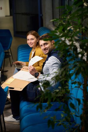 Téléchargez les photos : Collèges souriants travaillant avec des documents dans un espace de coworking sur un canapé bleu, femme dans une veste jaune - en image libre de droit