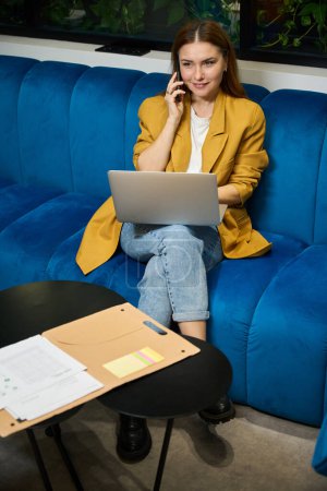 Téléchargez les photos : Belle jeune femme utilise un téléphone portable et un ordinateur portable, elle est située dans un espace de coworking sur un canapé confortable - en image libre de droit