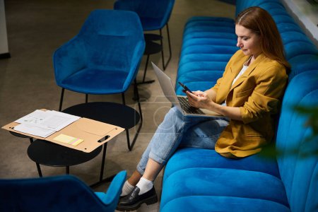 Foto de Mujer joven utiliza un teléfono móvil y un ordenador portátil, que se encuentra en un espacio de coworking en un cómodo sofá - Imagen libre de derechos