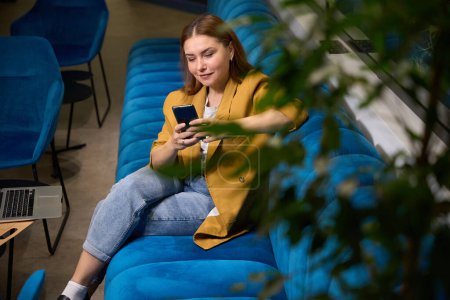 Foto de Mujer gerente se sienta con un teléfono en sus manos en un espacio de coworking en un cómodo sofá - Imagen libre de derechos