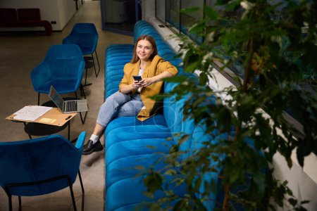 Foto de La freelancer se sienta con un teléfono en sus manos en un espacio de coworking en un cómodo sofá - Imagen libre de derechos