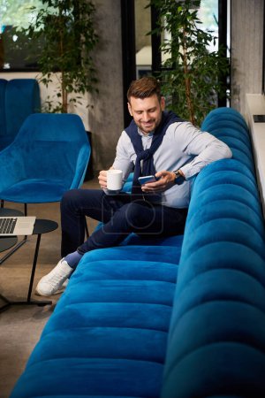 Téléchargez les photos : Gérant avec un téléphone portable et une tasse de café dans un espace de coworking, il s'assoit sur un canapé confortable - en image libre de droit