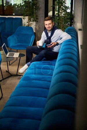Téléchargez les photos : Homme avec un téléphone portable et une tasse de café dans un espace de coworking, il est assis sur un canapé bleu - en image libre de droit