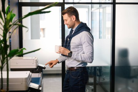 Téléchargez les photos : L'homme imprime des documents de travail sur l'équipement de bureau, il est dans des vêtements décontractés confortables - en image libre de droit