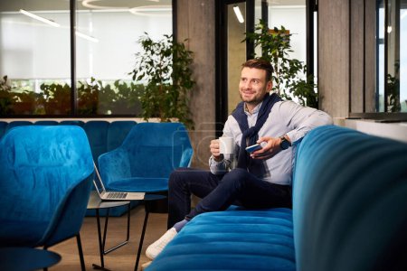 Téléchargez les photos : Jeune homme avec téléphone portable et une tasse de café dans un espace de coworking, il s'assoit sur un canapé bleu - en image libre de droit