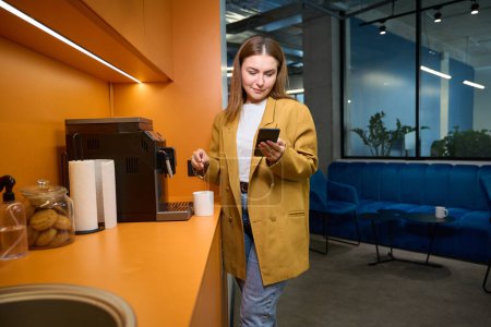 Téléchargez les photos : Femme freelance fait du thé dans la cuisine d'un espace de coworking, elle communique sur son téléphone portable - en image libre de droit