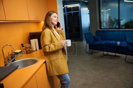 Téléchargez les photos : Femme avec une tasse de thé communique au téléphone dans la cuisine d'un espace de coworking - en image libre de droit