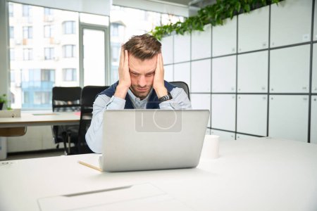 Photo for Man sits in front of a laptop at his workplace, he suffers from a headache - Royalty Free Image