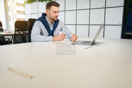 Téléchargez les photos : L'homme fatigué est assis sur son lieu de travail avec une tablette et un verre d'eau, ordinateur portable devant lui - en image libre de droit