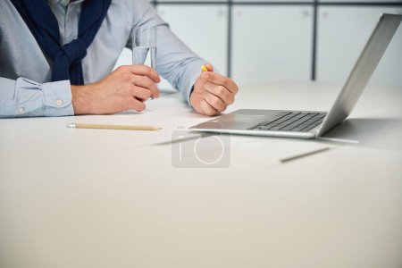 Téléchargez les photos : L'homme est assis sur son lieu de travail avec une tablette et un verre d'eau, un ordinateur portable devant lui - en image libre de droit