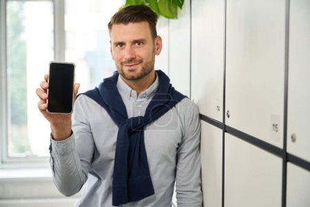 Téléchargez les photos : Jeune manager avec un téléphone portable se tient dans les casiers de stockage dans la zone de coworking bureau - en image libre de droit