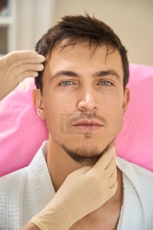Foto de Especialista cosmetólogo examina la cara de un hombre de pelo castaño, el cliente está sentado en una silla de cosmetología - Imagen libre de derechos