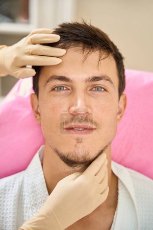 Foto de Especialista esteticista examina la cara de un hombre de pelo castaño, el cliente está sentado en una silla de cosmetología - Imagen libre de derechos
