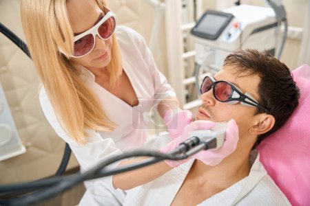 Photo for Blonde woman performs laser hair removal procedure on a young mans face, people wearing safety glasses - Royalty Free Image