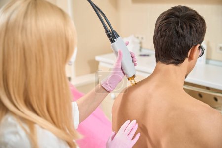 Photo for Blonde woman conducts session of laser hair removal on the shoulders of a young man, a professional uses modern equipment - Royalty Free Image