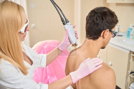 Téléchargez les photos : Cosmetologue effectue une séance d'épilation au laser des épaules d'un jeune homme, un professionnel utilise un équipement moderne - en image libre de droit