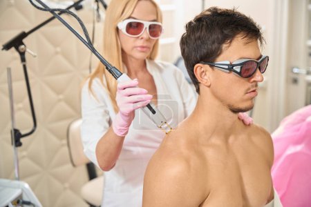 Téléchargez les photos : Femme cosmétologue conduit une séance d'épilation au laser des épaules de jeunes hommes, une femme blonde utilise un équipement moderne - en image libre de droit