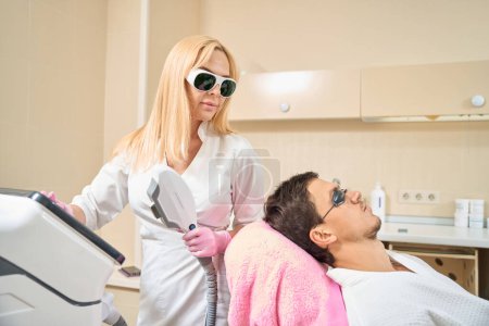 Foto de Cosmetólogo femenino ajusta el dispositivo para el procedimiento de elevación smas, el cliente está sentado en una silla de cosmetología - Imagen libre de derechos