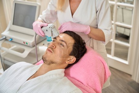 Téléchargez les photos : Docteur effectue une procédure de quincaillerie cosmétologique sur un client dans la région du front, l'homme est assis dans une chaise de cosmétologie - en image libre de droit