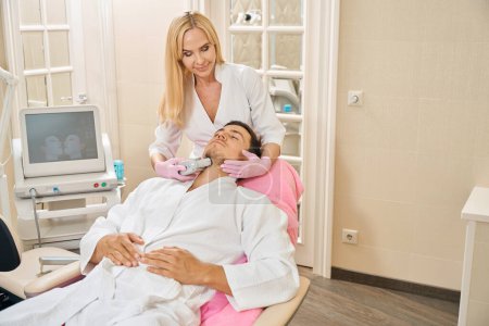 Foto de Mujer rubia realiza microneedle RF elevación en un cliente en el área de la barbilla, un hombre se sienta en una silla de cosmetología - Imagen libre de derechos