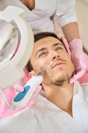 Foto de Hombre joven sometido a limpieza facial ultrasónica por hardware, un especialista que utiliza un dispositivo moderno - Imagen libre de derechos