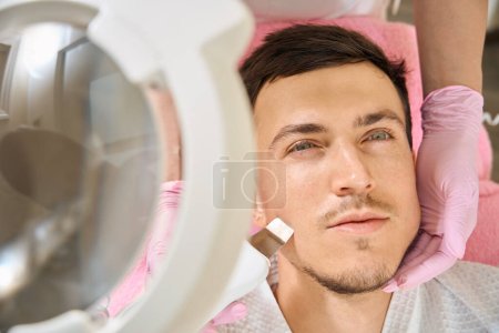 Téléchargez les photos : Jeune homme subissant matériel nettoyage du visage par ultrasons, le spécialiste utilise un dispositif moderne - en image libre de droit