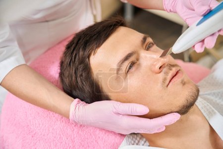 Téléchargez les photos : Masculin sur le nettoyage du visage par ultrasons matériel, spécialiste utilise un dispositif moderne - en image libre de droit