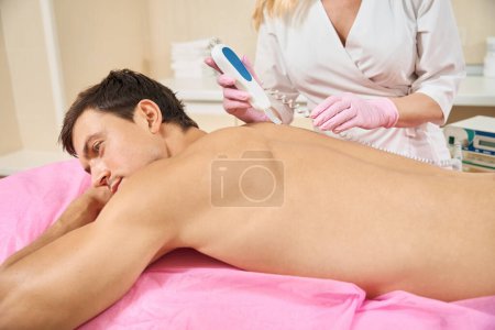Téléchargez les photos : L'homme au dos nu s'allonge sur un canapé de cosmétologie, le médecin procède à un nettoyage du matériel de sa peau du dos - en image libre de droit