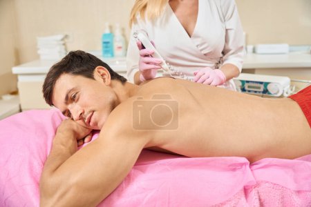 Téléchargez les photos : Jeune homme avec le dos nu se couche sur un canapé de cosmétologie, le médecin fait le nettoyage du matériel de sa peau du dos - en image libre de droit