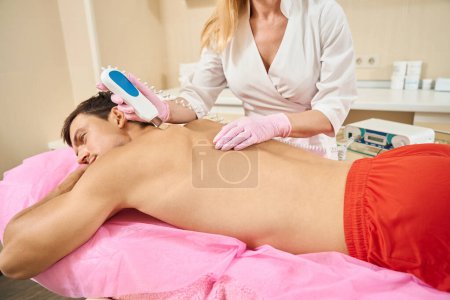 Photo for Cosmetologist does ultrasonic cleaning of the back skin of a young man, the specialist works in protective gloves - Royalty Free Image