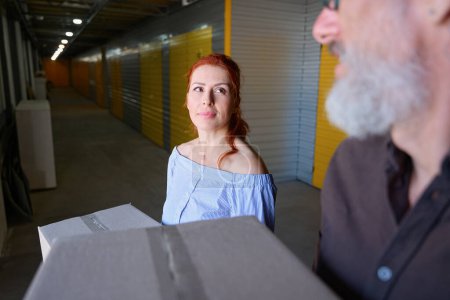 Téléchargez les photos : Femme d'âge moyen aux cheveux roux est dans un entrepôt de stockage avec son compagnon, les gens ont des boîtes en carton avec des choses - en image libre de droit