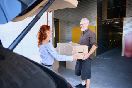 Photo for Bearded man and his wife are unloading the trunk of a car, they are carrying boxes with things - Royalty Free Image
