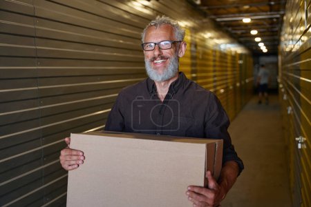 Téléchargez les photos : Homme souriant avec des lunettes porte une boîte en carton, il est dans un entrepôt - en image libre de droit
