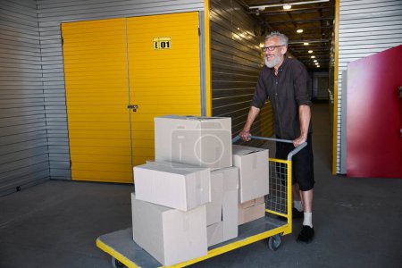 Photo for Cheerful man is carrying a lot of boxes on a cargo cart, he is in a warehouse - Royalty Free Image