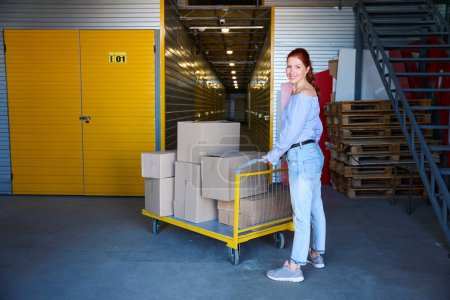 Foto de Mujer pelirroja se para con un carro de carga en un almacén, hay un montón de cajas de cartón en el carro - Imagen libre de derechos