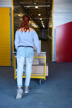 Foto de Mujer está empujando un carro de carga alrededor de un almacén, hay un montón de cajas de cartón en el carro - Imagen libre de derechos