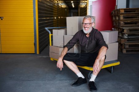 Photo for Cheerful man sat down to rest on a cart with boxes, he is in a warehouse - Royalty Free Image