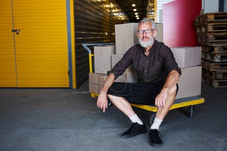 Téléchargez les photos : Homme à lunettes assis sur un chariot avec des boîtes, il est dans un entrepôt - en image libre de droit