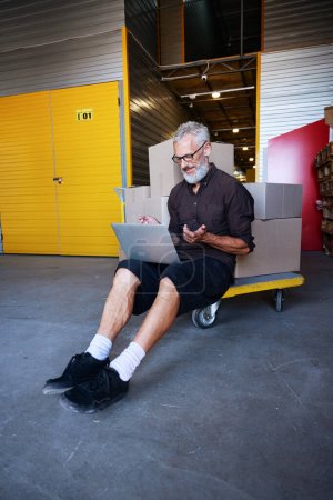 Téléchargez les photos : Homme d'âge moyen assis sur un chariot avec des boîtes, il communique sur un ordinateur portable - en image libre de droit