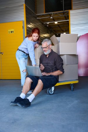 Photo for 35 plus couple is in a storage warehouse, the man is located with a laptop on a cargo cart with boxes - Royalty Free Image