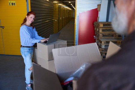 Photo for Nice woman using a laptop in a warehouse, next to a gray-haired man with boxes - Royalty Free Image
