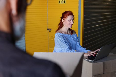 Foto de Mujer bonita está usando un portátil en un almacén, al lado de un hombre de pelo gris con cajas - Imagen libre de derechos