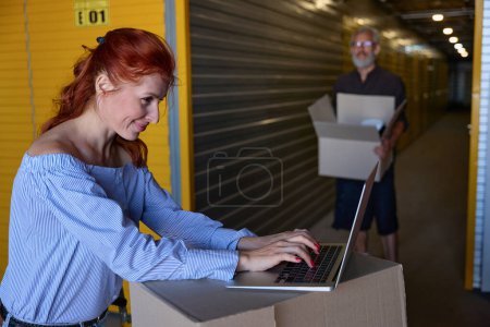 Foto de Alegre pareja 35 plus se comunica en un almacén de almacenamiento, la mujer utiliza un ordenador portátil - Imagen libre de derechos