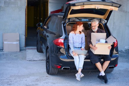 Téléchargez les photos : Couple assis sur le bord du coffre ouvert, ils ont apporté des choses à l'entrepôt en voiture - en image libre de droit