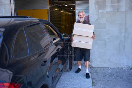 Téléchargez les photos : L'homme barbu porte des boîtes d'une voiture, il est dans un entrepôt - en image libre de droit