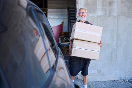 Téléchargez les photos : Un homme aux cheveux gris porte des boîtes d'une voiture, il est dans un entrepôt - en image libre de droit
