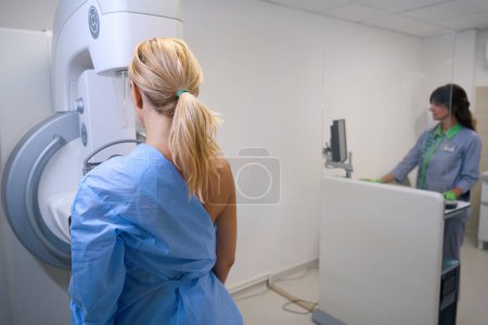 Erwachsene Frau steht vor der Mammographie-Einheit, die vom Röntgen über die Bedienkonsole im Hintergrund bedient wird